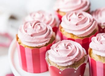 Strawberry cupcakes with strawberry buttercream, in pink, red, and white cupcake papers, sitting on a white cake stand. The buttercream has been piped in the shape of a rosette, and there is a pink-trimmed cloth in the background.