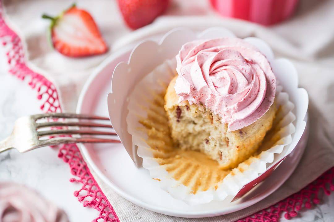 Strawberry cupcakes with strawberry buttercream, in pink, red, and white cupcake papers, sitting on a white cake stand. The buttercream has been piped in the shape of a rosette, and there is a pink-trimmed cloth in the background.