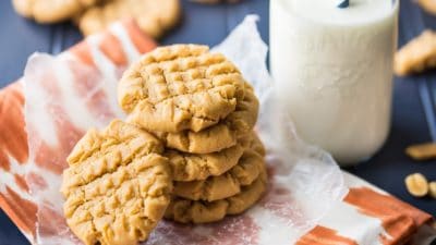A stack of peanut butter cookies on a napkin, with a bottle of milk in the background. Cookies are placed on an orange napkin, with a dark blue tabletop below.