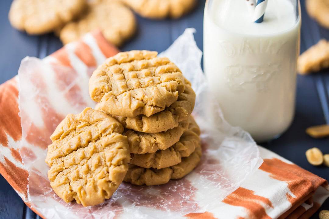 A stack of peanut butter cookies on a napkin, with a bottle of milk in the background.  Cookies are placed on an orange napkin, with a dark blue tabletop below.