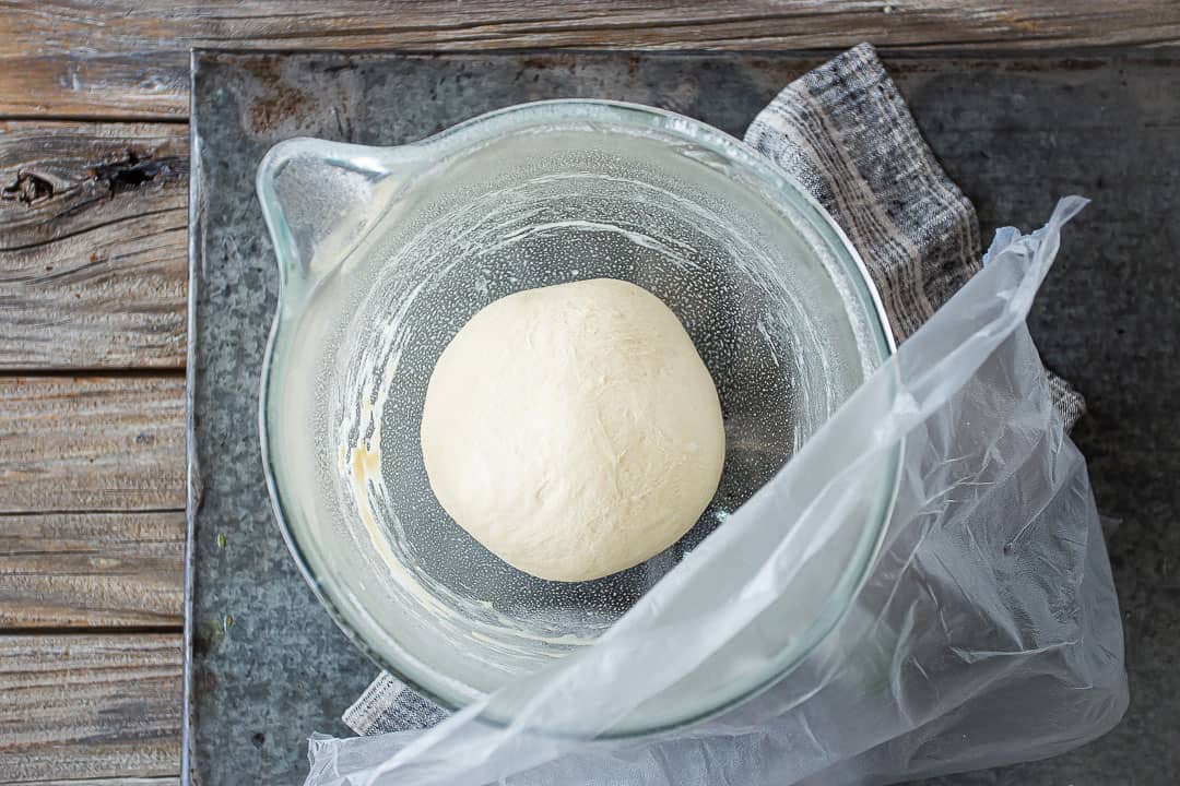Resting pizza dough recipe in a large oiled bowl with plastic wrap on top.