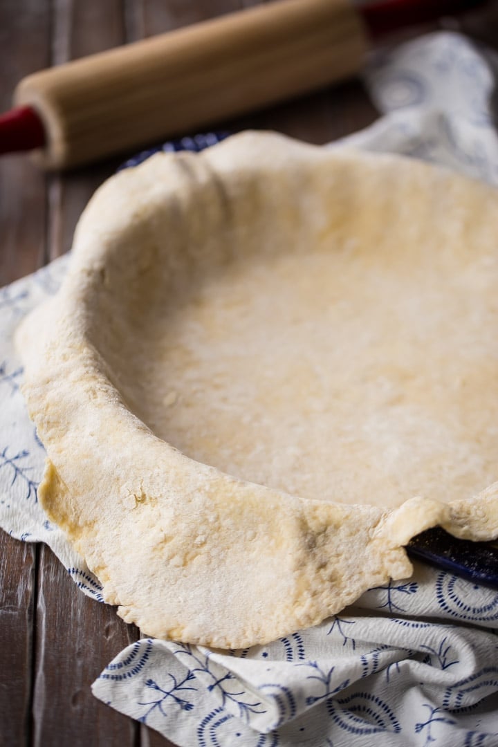 Vertical image of homemade pie dough draped over a pie pan, with a rolling pin in the background.