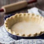 Square image of unbaked pie crust in a dark blue dish with a rolling pin in the background.