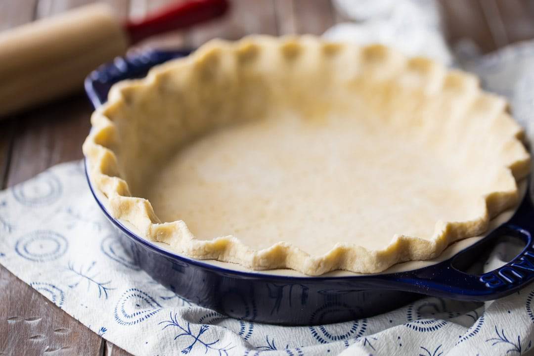 Horizontal image of unbaked pie crust in a dark blue dish with a rolling pin in the background.