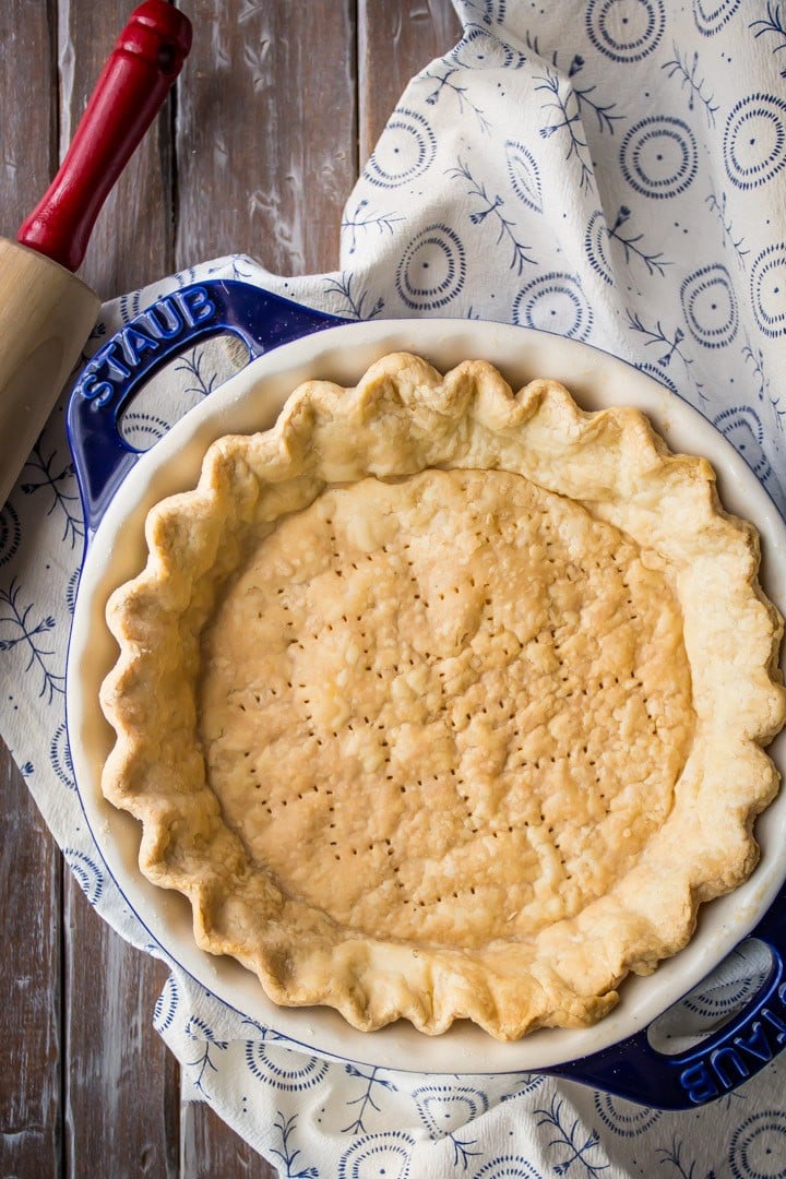 Vertical overhead image of flaky pie crust baked in a blue dish with a blue printed cloth and a rolling pin.