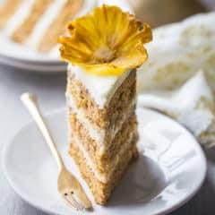 A slice of hummingbird cake with cream cheese frosting and a dried pineapple flower on a white plate.