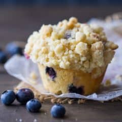 Square image of a bakery-style blueberry muffin with streusel crumb topping on a dark wood background with fresh blueberries scattered around.