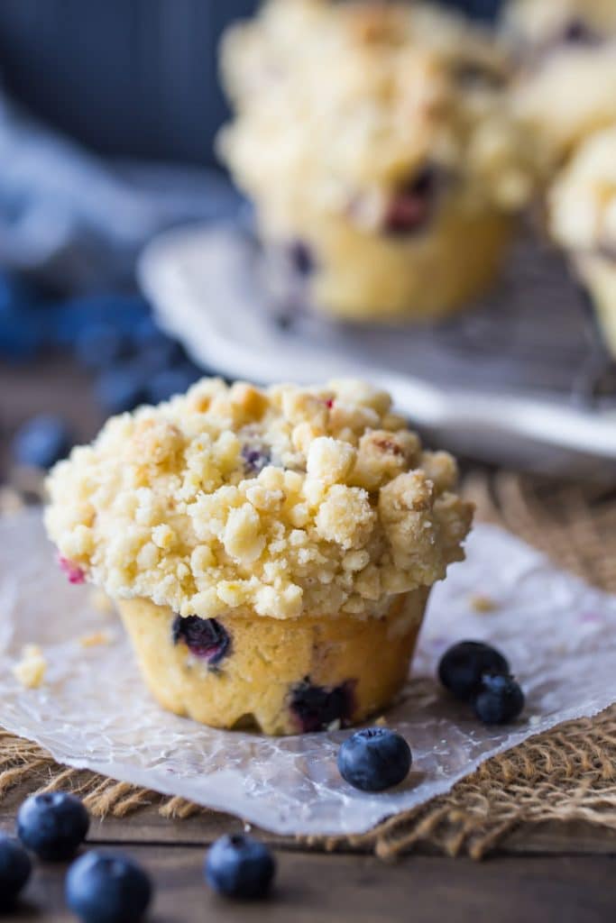 Vertical image of a crumble-topped blueberry muffin on a dark background, with a tray of blueberry muffins behind.