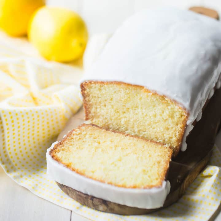 Iced lemon loaf on a wood cutting board with lemons in the background.