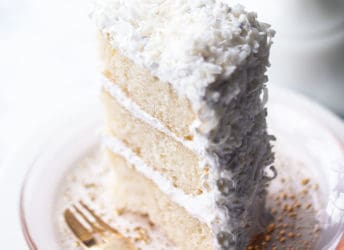 Tall slice of coconut cake on a pink plate with a gold fork.