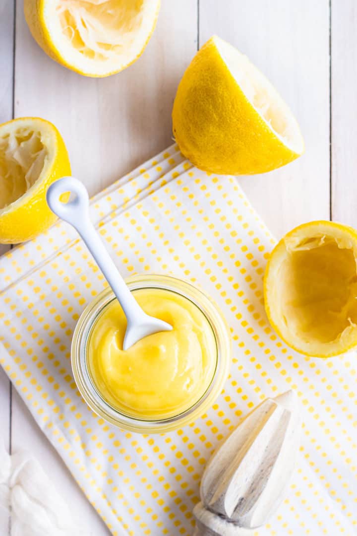 Overhead image of a jar of lemon curd surrounded by juiced lemons, with a citrus reamer and a yellow checked cloth.