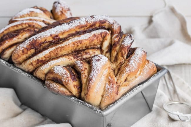 A loaf of braided bread is baked in a rectangular metal pan. The bread is golden brown with dark cinnamon swirls and topped with a light dusting of powdered sugar. A light fabric napkin is loosely draped underneath the pan on a white surface.