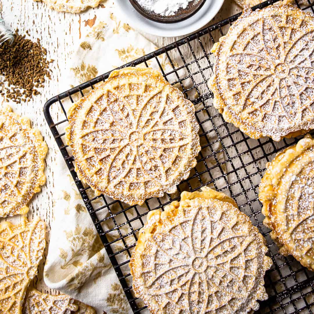 Overhead image of pizzelle on a wire cooling rack with whole anise seeds and powdered sugar.