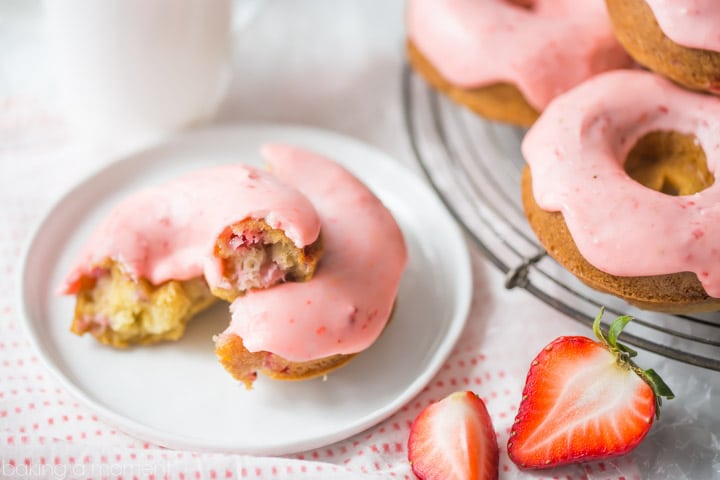 Baked strawberry donut, broken in half on a plate, with fresh strawberry and a stack of donuts on a cooling rack.
