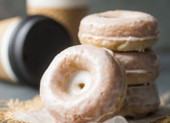 Stacks of homemade glazed sour cream donuts, with coffee cups in the background.