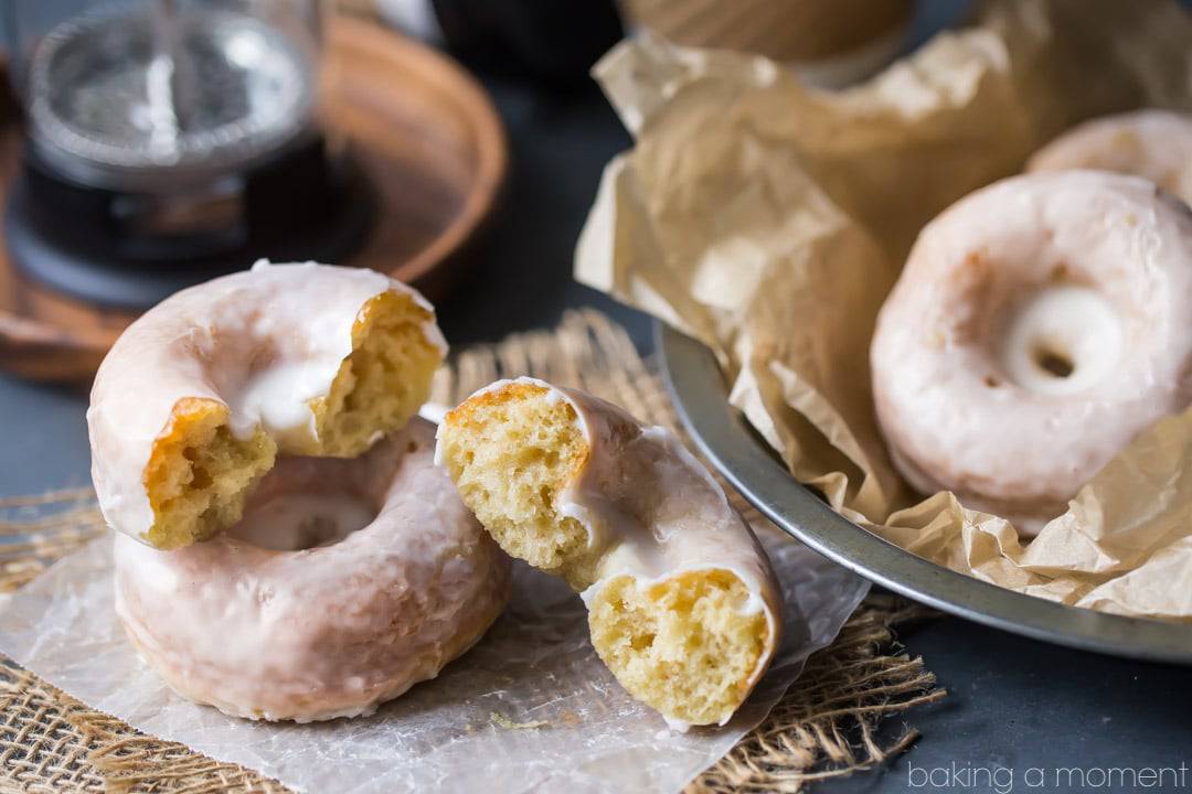 Glazed sour cream cake donuts, with one broken in half.  Coffee pot and coffee cups in the background. 