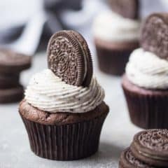 Chocolate Cupcake with Cookies and Cream Frosting and an Oreo Cookie on top, with more cupcakes in the background and stacks of cookies scattered around.