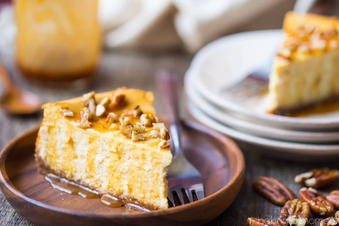 Caramel pecan cheesecake slice on a plate, garnished with caramel sauce and chopped pecans, with a stack of plates and a jar of caramel sauce in the background