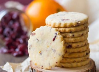 Stack of cranberry orange shortbread cookies on a wooden coaster, with a jar of dried cranberries and a whole orange in the background.