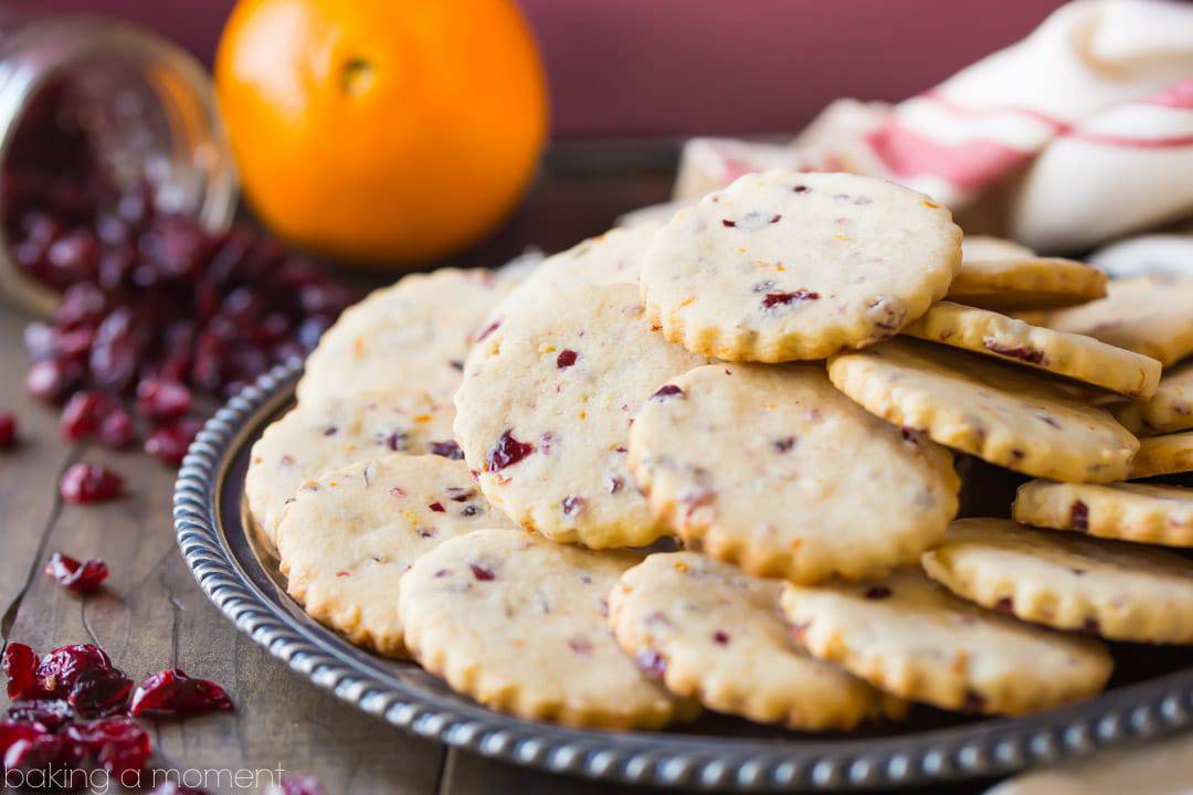 Platter of cranberry orange shortbread cookies, in front of a maroon background with whole orange and dried cranberries in the background. 
