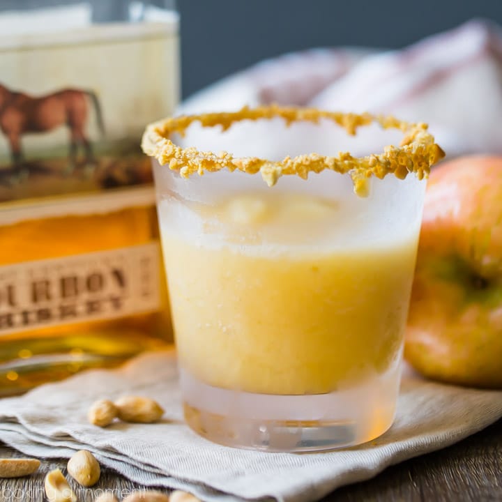 Caramel apple slush cocktail in a rocks glass with caramel and crushed peanuts around the rim.  Fresh apple and a bottle of bourbon in the background, with peanuts scattered in the foreground. 
