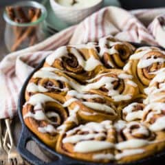 Cast iron skillet with gingerbread cinnamon rolls, drizzled with cream cheese icing. Cinnamon sticks and gold forks in the background.