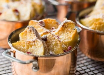 Individual bread & butter puddings, in mini copper pots, with toasty buttered bread, dried cranberries, and powdered sugar, on a wire cooling rack.