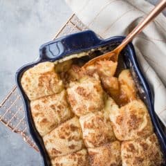 Snickerdoodle Apple Cobbler in a dark blue casserole dish: apple pie filling topped with cinnamon sugar snickerdoodle cookies, on a gray background with a copper serving spoon.