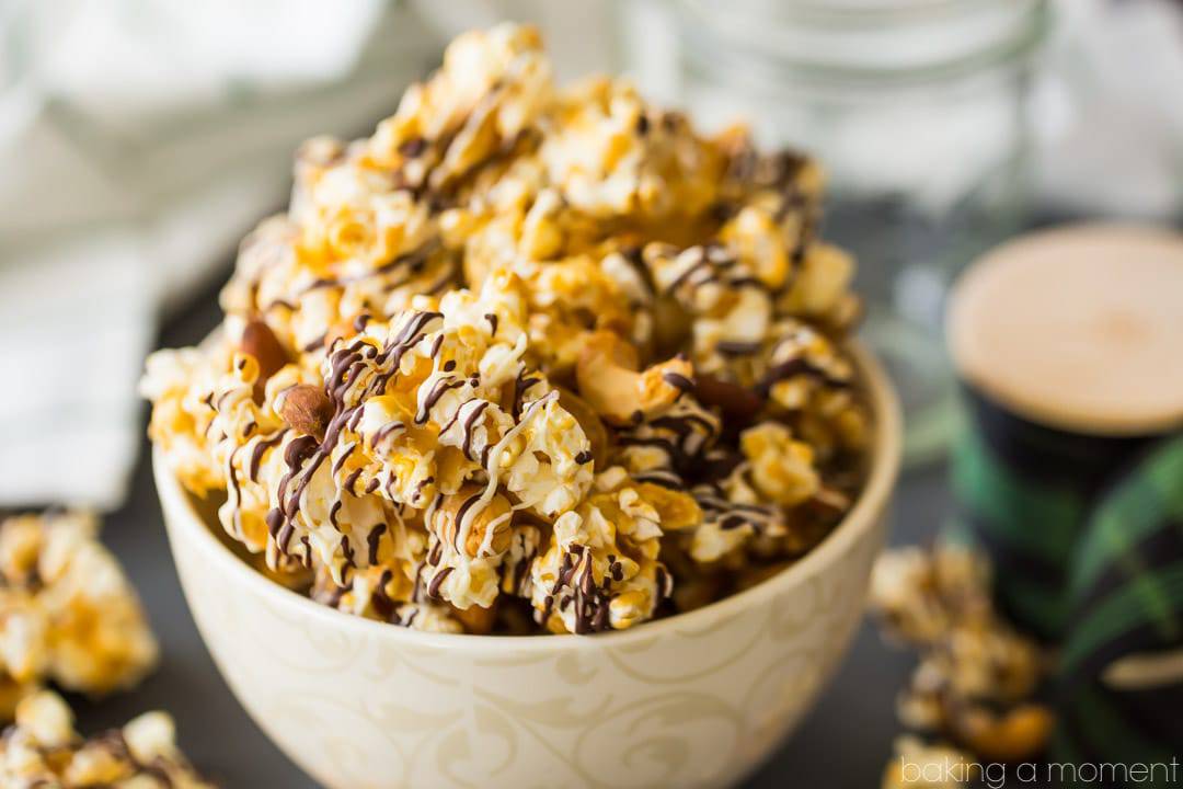 Homemade Moose Munch butter toffee caramel popcorn with nuts and chocolate drizzle, in a bowl with a spool of holiday ribbon in the background
