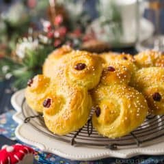 Saffron yellow St. Lucia Buns in an "S" shape, sprinkled with sugar and garnished with dried cranberries, on a cooling rack with a dark blue background with Christmas decorations.