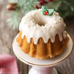 Mini brown sugar pound cake bundt on a white stand with white icing, white sprinkles, and a sprig of royal icing holly leaves & berries, with holiday greenery and a red checked napkin in the background.