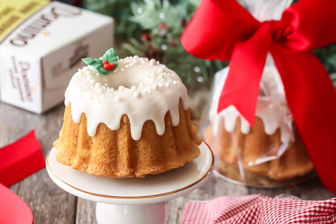 Mini brown sugar pound cake bundt on a white stand with white icing, white sprinkles, and a sprig of royal icing holly leaves & berries, with holiday greenery and a red checked napkin in the background. 
