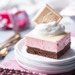 Chocolate Strawberry Mousse Bar: a layered dessert with chocolate on the bottom, and a pale pink strawberry mousse drizzled with white chocolate glaze. Topped with whipped cream and a Ghirardelli Strawberry Bark SQUARE, plated and sitting on a pink napkin with gold forks and candy wrappers in the background.