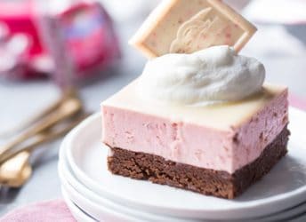 Chocolate Strawberry Mousse Bar: a layered dessert with chocolate on the bottom, and a pale pink strawberry mousse drizzled with white chocolate glaze. Topped with whipped cream and a Ghirardelli Strawberry Bark SQUARE, plated and sitting on a pink napkin with gold forks and candy wrappers in the background.