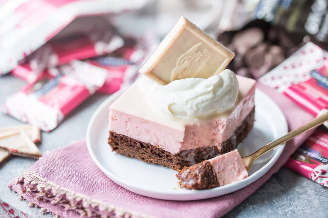 Chocolate Strawberry Mousse Bar: a layered dessert with chocolate on the bottom, and a pale pink strawberry mousse drizzled with white chocolate glaze.  Topped with whipped cream and a Ghirardelli Strawberry Bark SQUARE, plated and sitting on a pink napkin with gold forks and candy wrappers in the background. 