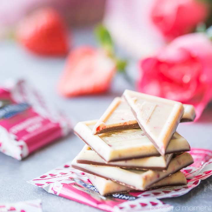 A stack of Ghirardelli Strawberry Bark SQUARES with roses, strawberries, and candy wrappers in the background. 