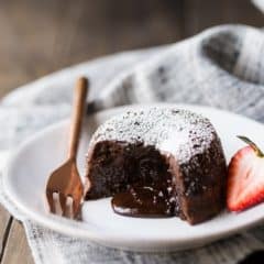 Chocolate Molten Lava Cake on a white plate, with liquid chocolate spilling from the center. Garnished with a strawberry, and a linen cloth beneath the plate and a copper fork on the side.