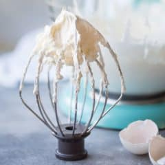 Whisk attachment with Swiss meringue buttercream and blue mixer in background with broken eggshells off to one side.