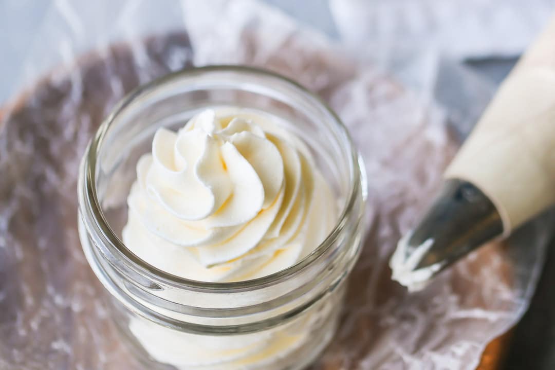 A small glass jar full of piped Swiss meringue buttercream, sitting on a wood plate with a piping bag off to one side.