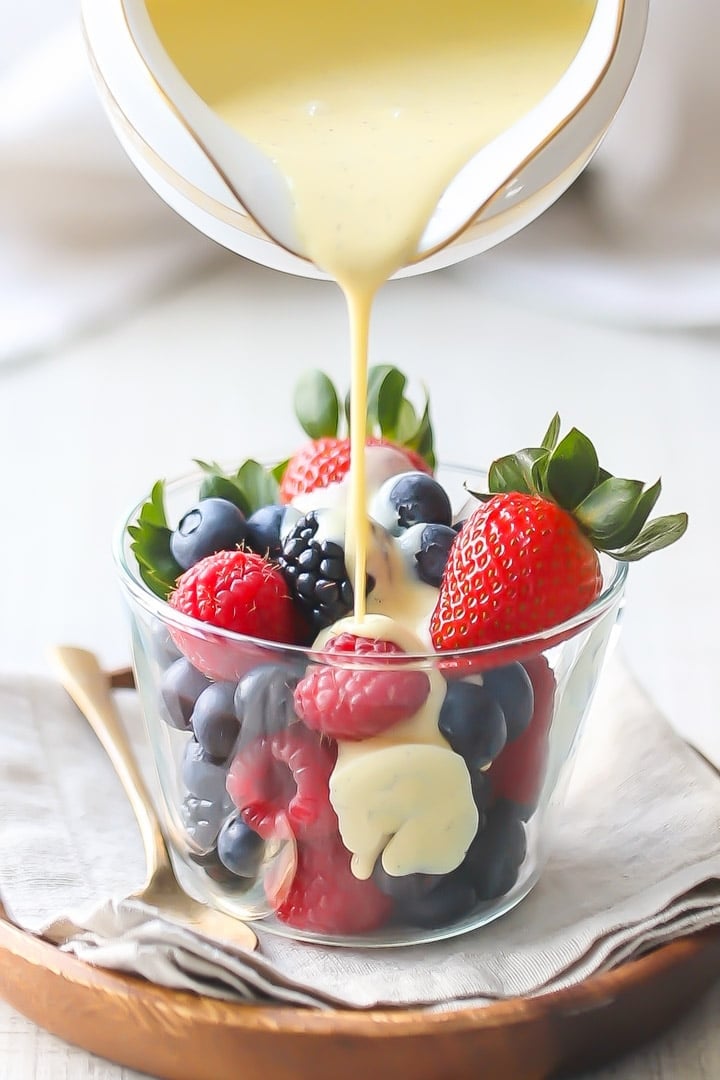 Creme Anglaise: rich, velvety vanilla custard sauce being poured over a bowl of berries, sitting on a linen napkin with a gold spoon off to one side.