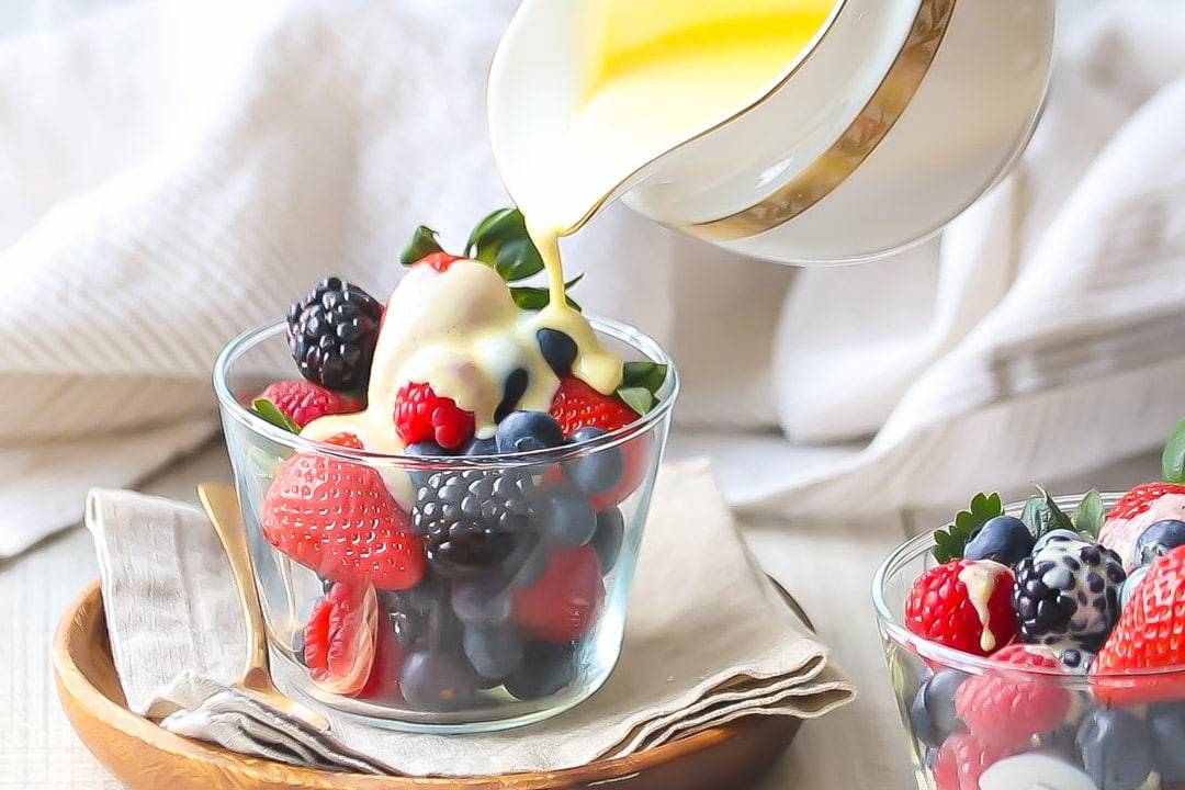 Creme Anglaise: rich, velvety vanilla custard sauce being poured over a bowl of berries, sitting on a linen napkin with a gold spoon off to one side.