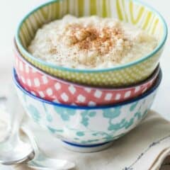 Three colorful bowls stacked on a linen napkin, filled with a creamy rice pudding and sprinkled with cinnamon.