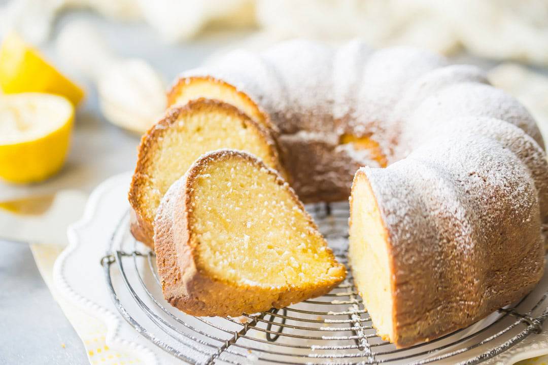 A moist lemon pound bundt cake, dusted with powdered sugar, on a wire cooling rack over a yellow napkin.