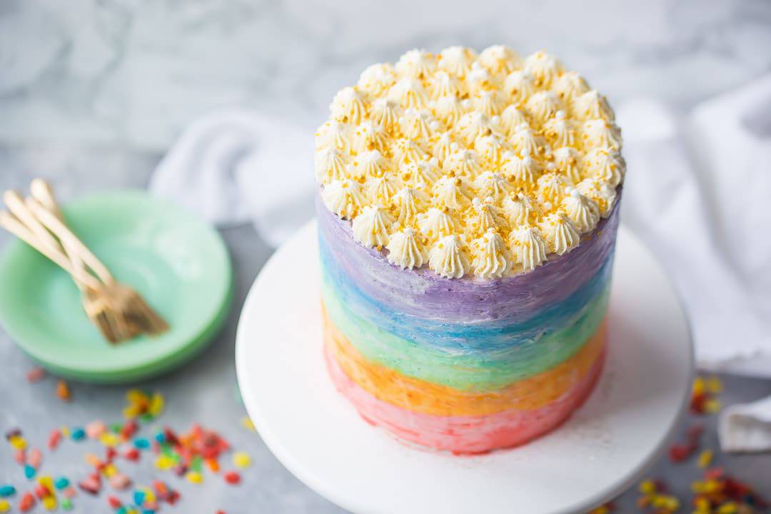 Rainbow fruity pebble cake with gold sprinkles on a cake stand with green plates, fruity pebbles cereal, and gold forks on a light gray background.  