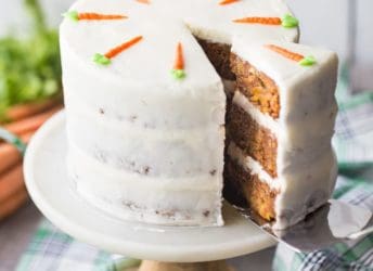 Cake server removing a slice of triple-layer carrot cake with cream cheese frosting, with a green plaid cloth below and fresh carrots in the background.