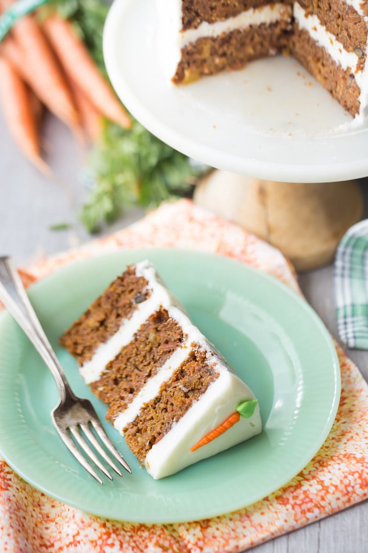 Slice of triple-layer carrot cake with cream cheese frosting, on a green plate with fresh carrots in the background