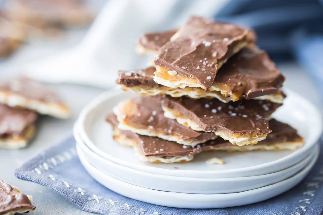 Horizontal image of a stack of matzo toffee on a white plate, with pieces of broken toffee in the background.