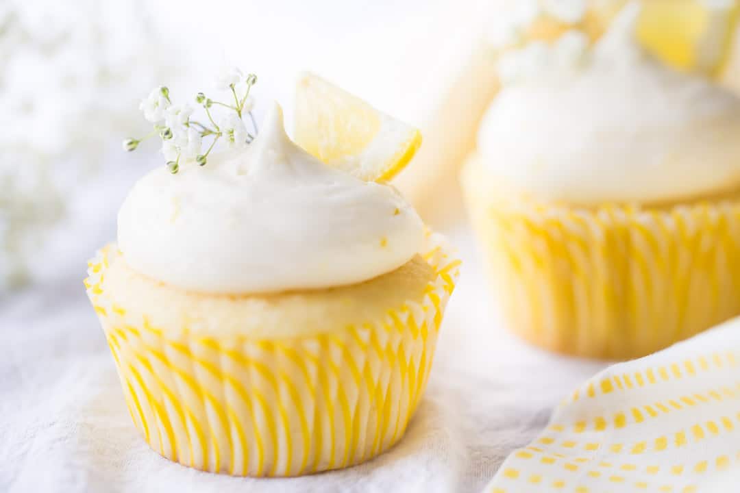 Horizontal image of lemon cupcakes with lemon curd filling & lemon cream cheese frosting, garnished with lemon wedges and a sprig of baby's breath.