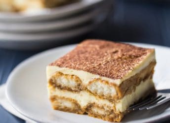 Square image of a slice of tiramisu on a white plate over a dark blue background.