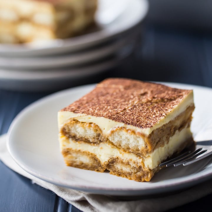 Square image of a slice of tiramisu on a white plate over a dark blue background.